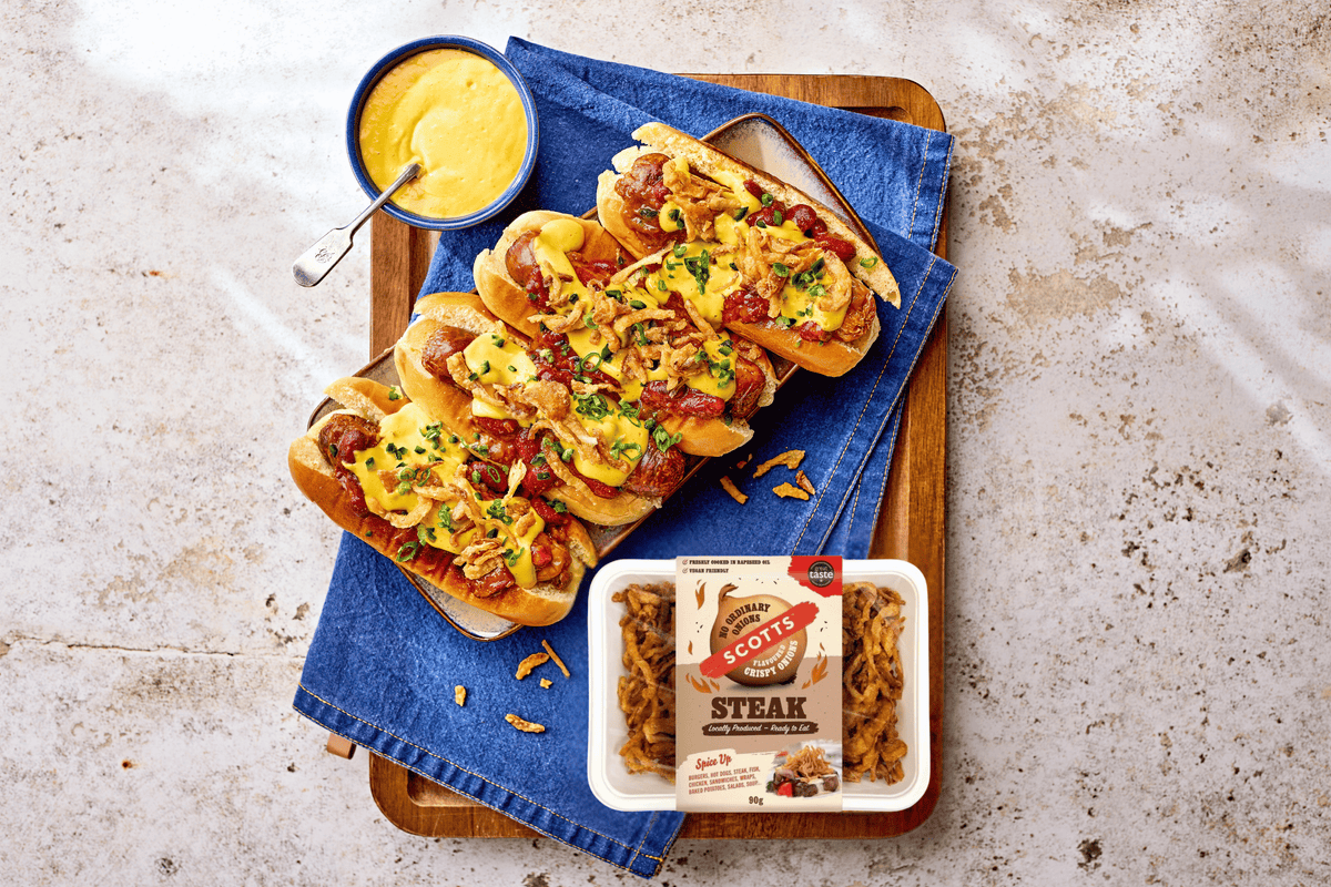 Four loaded hot dogs topped with melted cheese, bacon bits, and chopped green onions, arranged on a wooden board with a blue napkin. A small bowl of yellow mustard sauce sits at the top, and a package of Scott's Steak Fries is placed at the bottom right on a light stone surface.